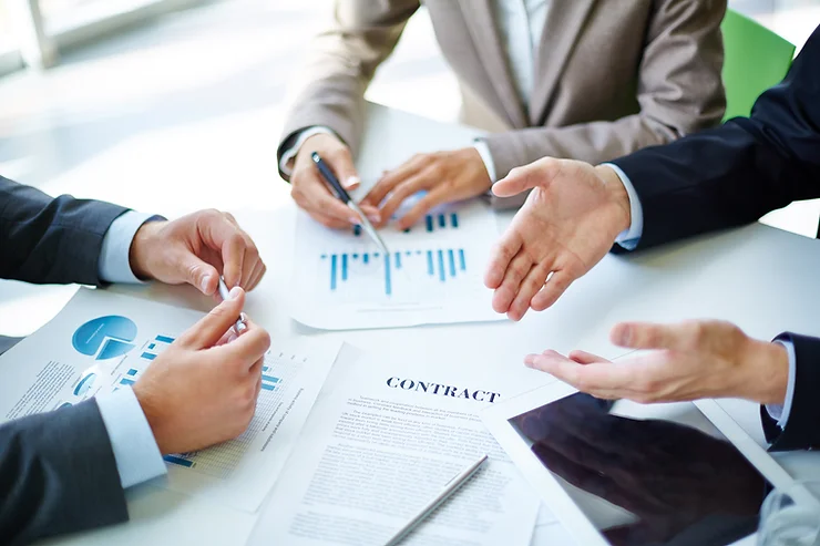 Three business people discuss charts and a contract at a table.