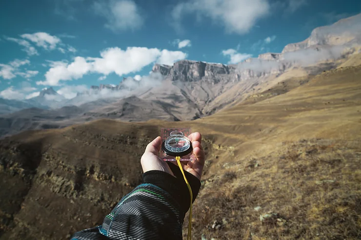 Person holding a compass against a vast mountainous landscape under a partly cloudy sky.