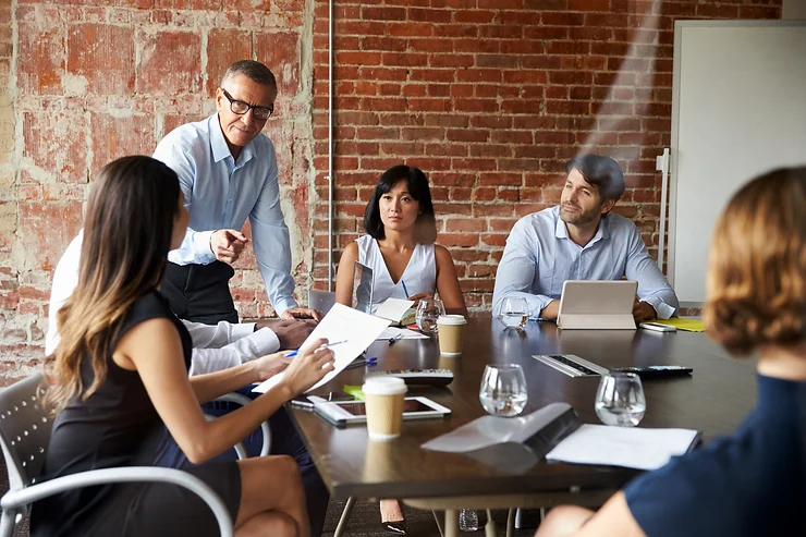 A group of people in a meeting room with a brick wall, discussing around a table with laptops, documents, and drinks. A man is standing and pointing while others sit and listen.
