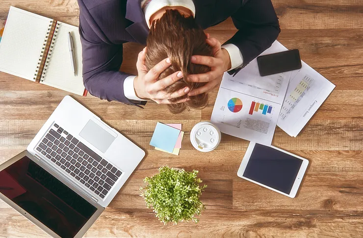 Person in business attire sitting at a cluttered desk with a laptop, documents, coffee, and a tablet, holding their head in frustration.