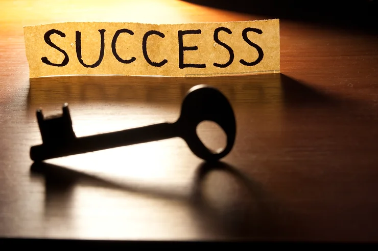 A black key lies on a surface in front of a strip of paper with the word "SUCCESS" written on it, lit by warm light.