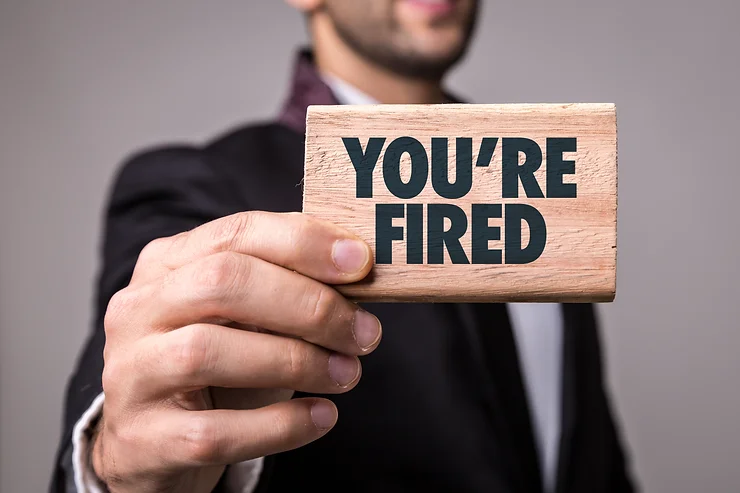 A man in a suit holds a wooden sign with the text "YOU'RE FIRED.