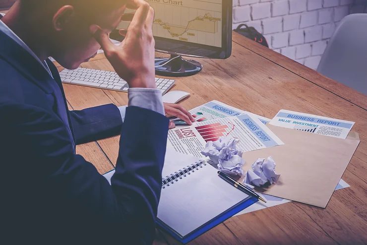 A person in business attire looks stressed, sitting at a desk with a computer, documents labeled "Business Report" and "Loss," and crumpled paper.