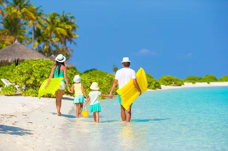 A family of four walks along a tropical beach, carrying yellow floats. They wear summer hats and are surrounded by clear blue water and lush greenery.