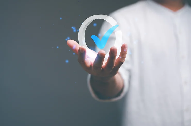 A person wearing a white shirt holds a glowing blue checkmark in an open hand against a dark background.