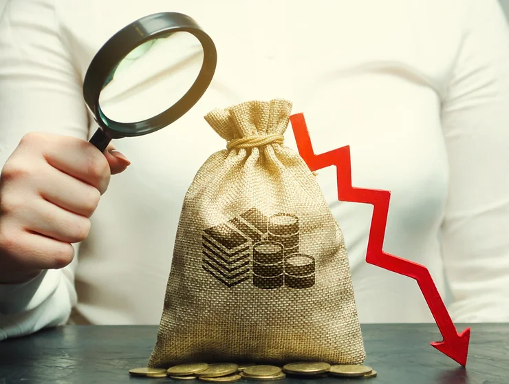 A person holds a magnifying glass over a burlap sack with coins, next to a downward red arrow on a table.
