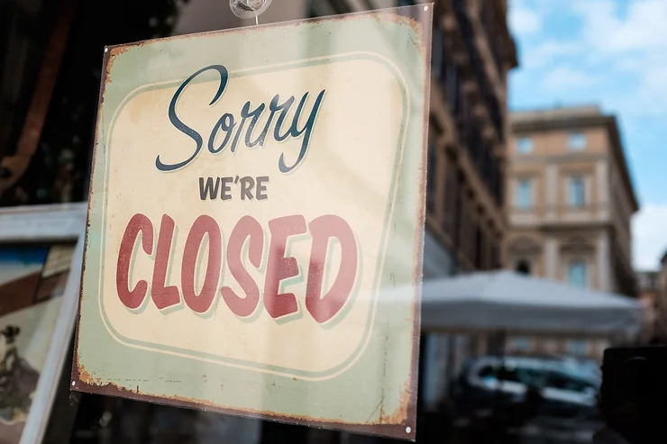 Sign on a storefront window reads "Sorry We're Closed" in decorative font, with a blurred street and buildings visible in the background.