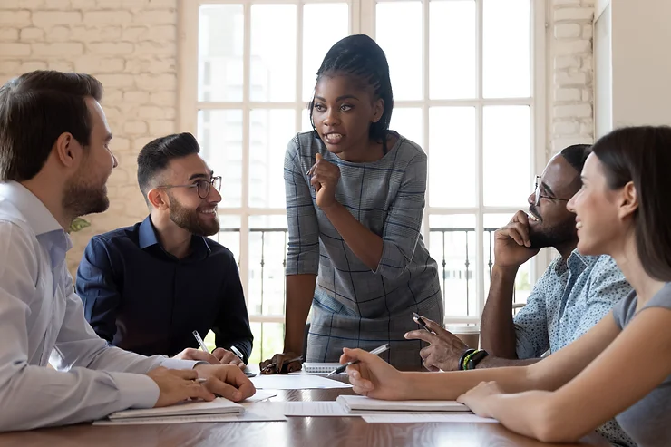 A group of people in a meeting room; a woman is standing and speaking, while four others, two men and two women, are seated and listening attentively.