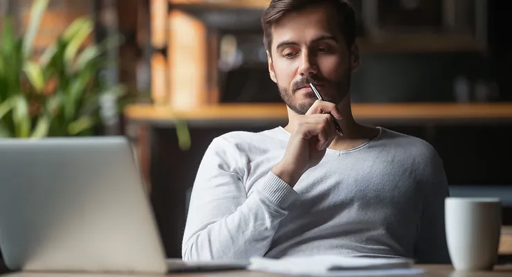 Man sitting at a table, holding a pen to his lips, looking at a laptop screen, with a mug nearby and greenery in the background.