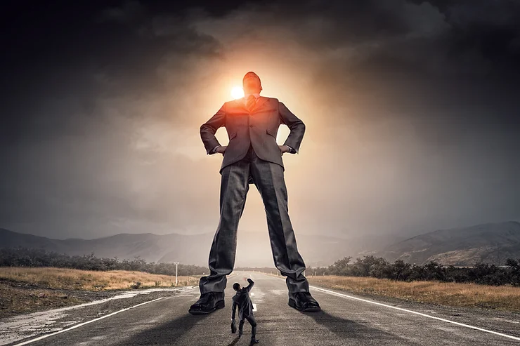 A small man faces a towering giant figure in a suit on a deserted road at sunset, with mountains in the background.