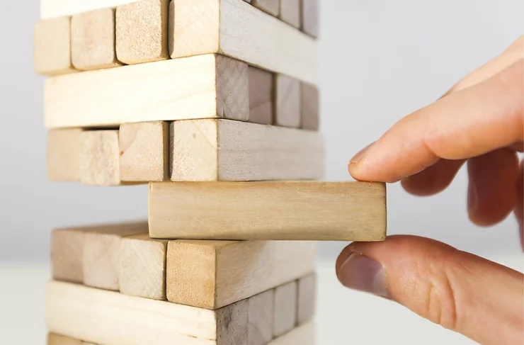 A hand is pulling a wooden block from a stacked Jenga tower, creating a potential risk of collapse.