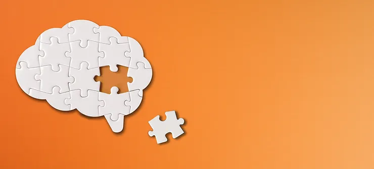 White jigsaw puzzle pieces assembled in the shape of a brain on an orange background, with one piece missing and placed nearby.