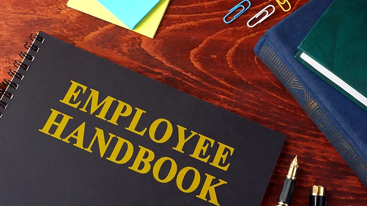 A spiral-bound employee handbook on a wooden desk with scattered paperclips, papers, and a pen nearby.
