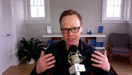 Person speaking into a microphone in a study with shelves and a plant in the background.
