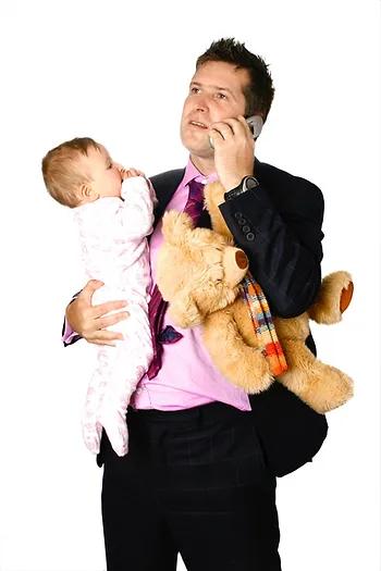 A man in a suit holds a baby and a teddy bear while talking on a cellphone.