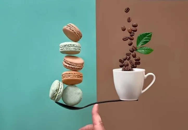 A spoon balanced on a finger holds four stacked macarons. Coffee beans and leaves rise from a cup nearby, set against a split teal and brown background.