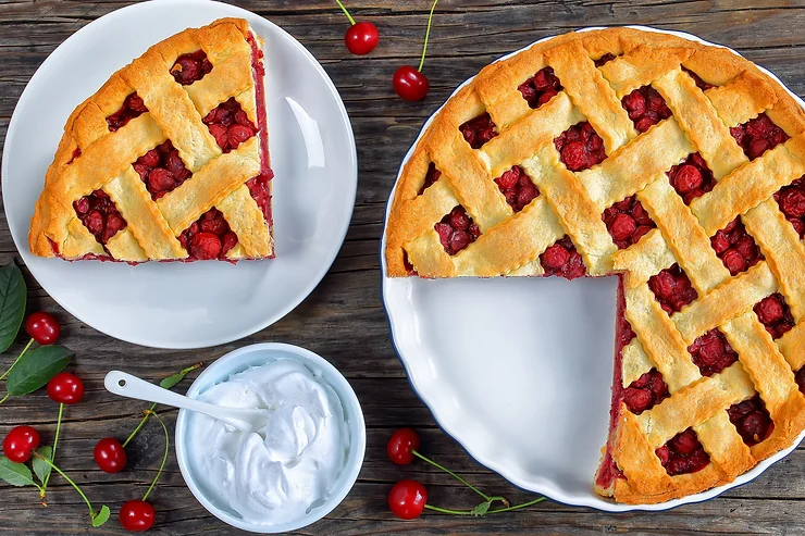 Cherry pie with lattice crust, partially sliced, on a wooden table next to a plate with a pie slice and a bowl of whipped cream.
