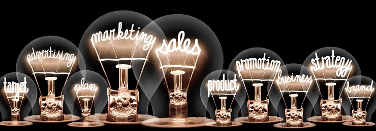 A row of lit light bulbs with words like target, advertising, marketing, sales, product, promotion, business, strategy, plan, and brand inside, against a black background.