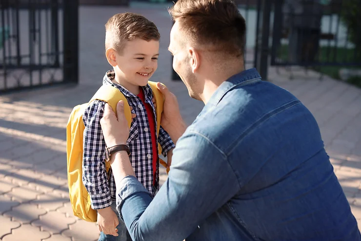 An adult kneels in front of a smiling child with a yellow backpack. They appear to be outside near a gate.