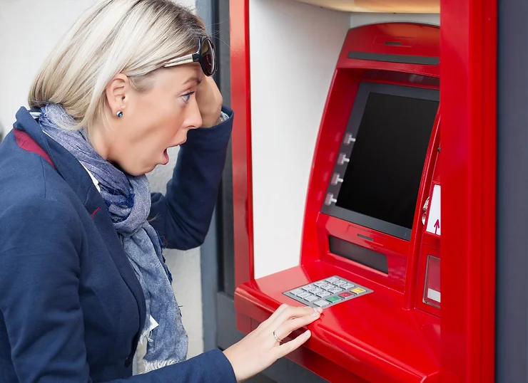 A shocked woman stands at a red ATM, looking at the screen with her mouth open in surprise.