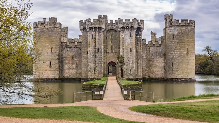 Medieval stone castle surrounded by a moat, featuring multiple cylindrical towers and an arched entrance, set against a cloudy sky.