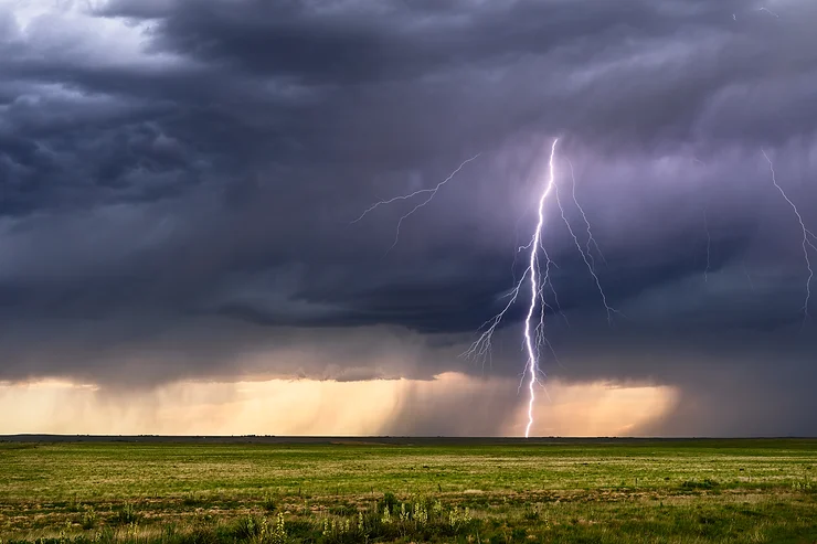 A bolt of lightning strikes a grassy open field under dark stormy clouds with rain in the distance.