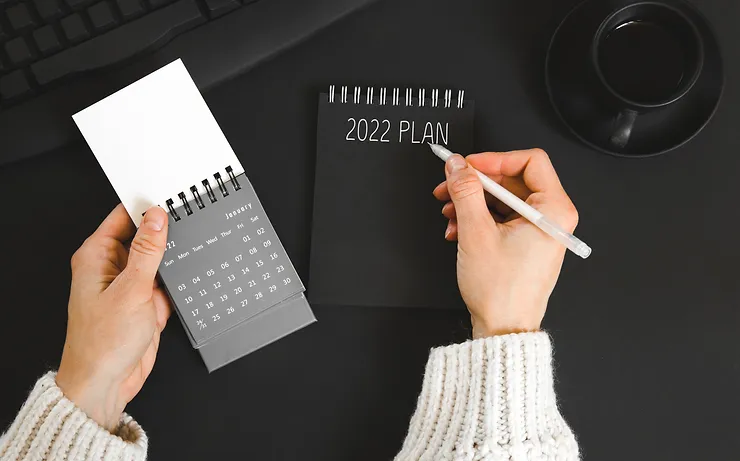 A person writes "2022 Plan" on a black notepad. A mini calendar and a cup of coffee are nearby on a black desk.