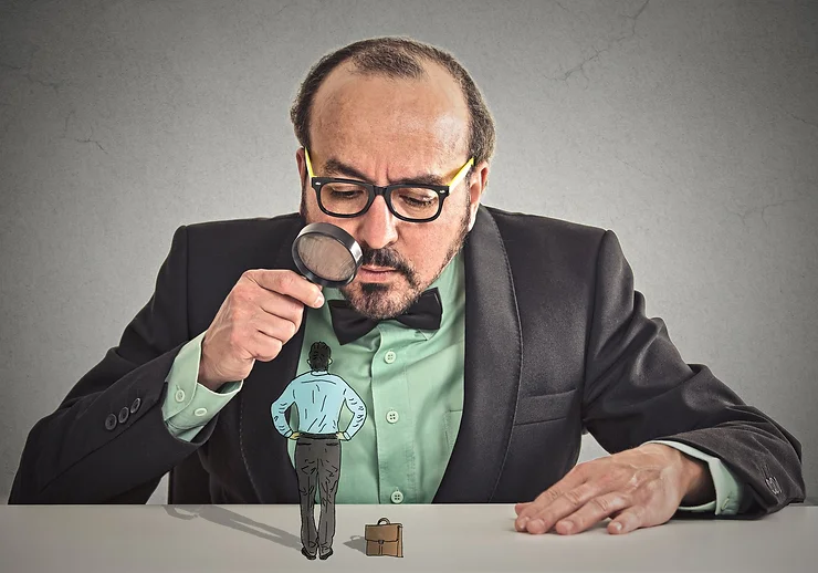 A man in a suit uses a magnifying glass to closely examine a miniature person standing on a table.