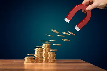 A hand holds a red magnet attracting stacks of gold coins from a wooden surface, set against a dark blue background.