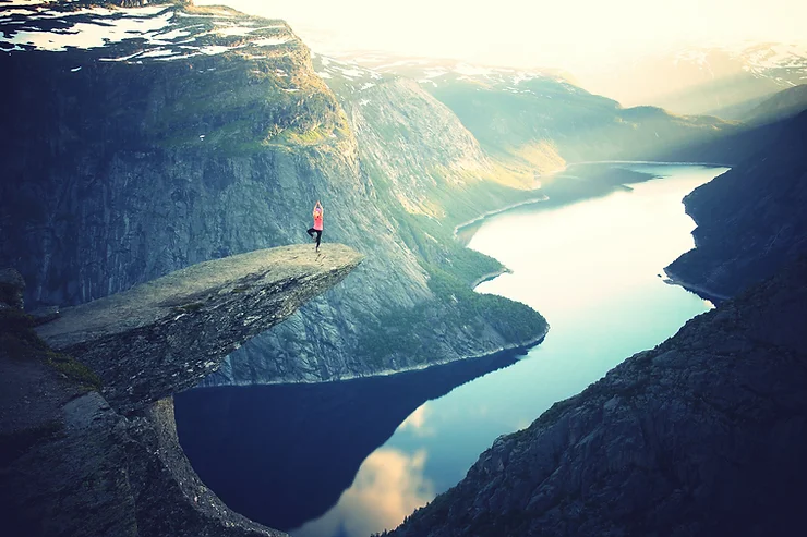 A person stands on a rocky outcrop overlooking a fjord with mountains in the background.