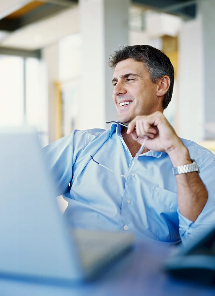 A man in a blue shirt smiles while sitting at a desk with a laptop, holding his glasses in one hand.