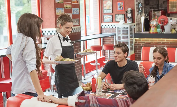A waitress serves food to a group of four friends sitting in a retro-style diner with red chairs and brick interiors.