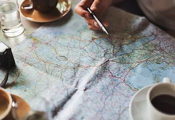 Hand holding a pen over a detailed map on a table with coffee cups.