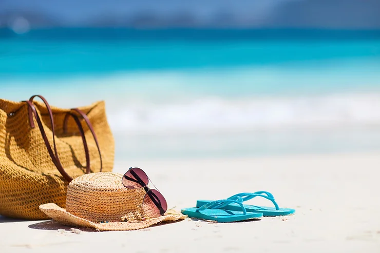 Straw bag and hat with sunglasses and blue flip-flops on a sandy beach, with turquoise ocean in the background.