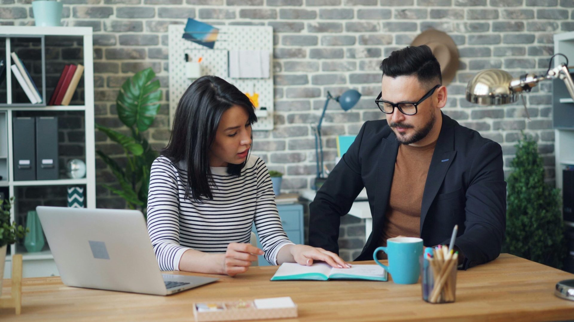 Two people sitting at a wooden desk with a laptop and mug, discussing notes in a blue folder. Brick wall and shelves in the background.